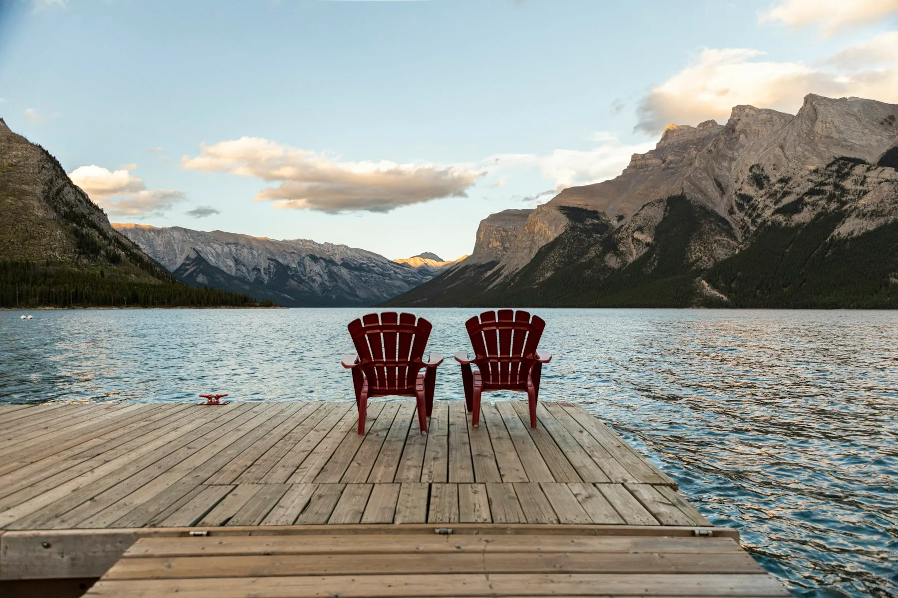 Two red Adirondack chairs on a dock facing a mountain lake